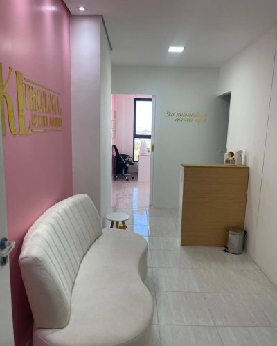 Sala de recepção da Clínica de Estética Dra. Karen Lima mostrando um sofá, onde os pacientes podem esperar confortavelmente pelo seu atendimento. Logo acima do sofá esta o logotipo da clínica KL Tricologia e Estética Avançada. Mostra tamém o balcão de atendimento e logo mais ao fundo a sala onde são feitos os procedimentos